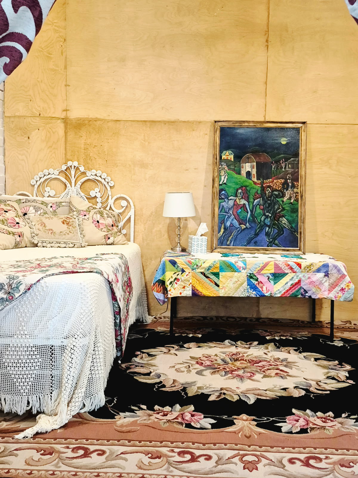 A bed dressed in crochet and appliqué linens beside a painted canvas and a lamp
