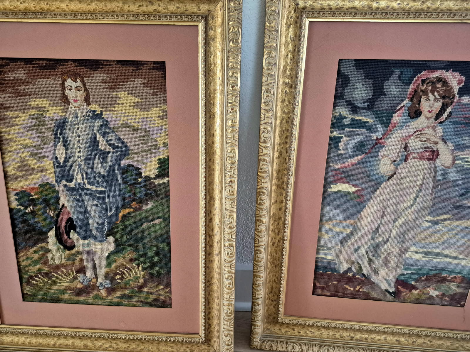 Two needlepoint portraits in gilt frames leaning against a pink wall