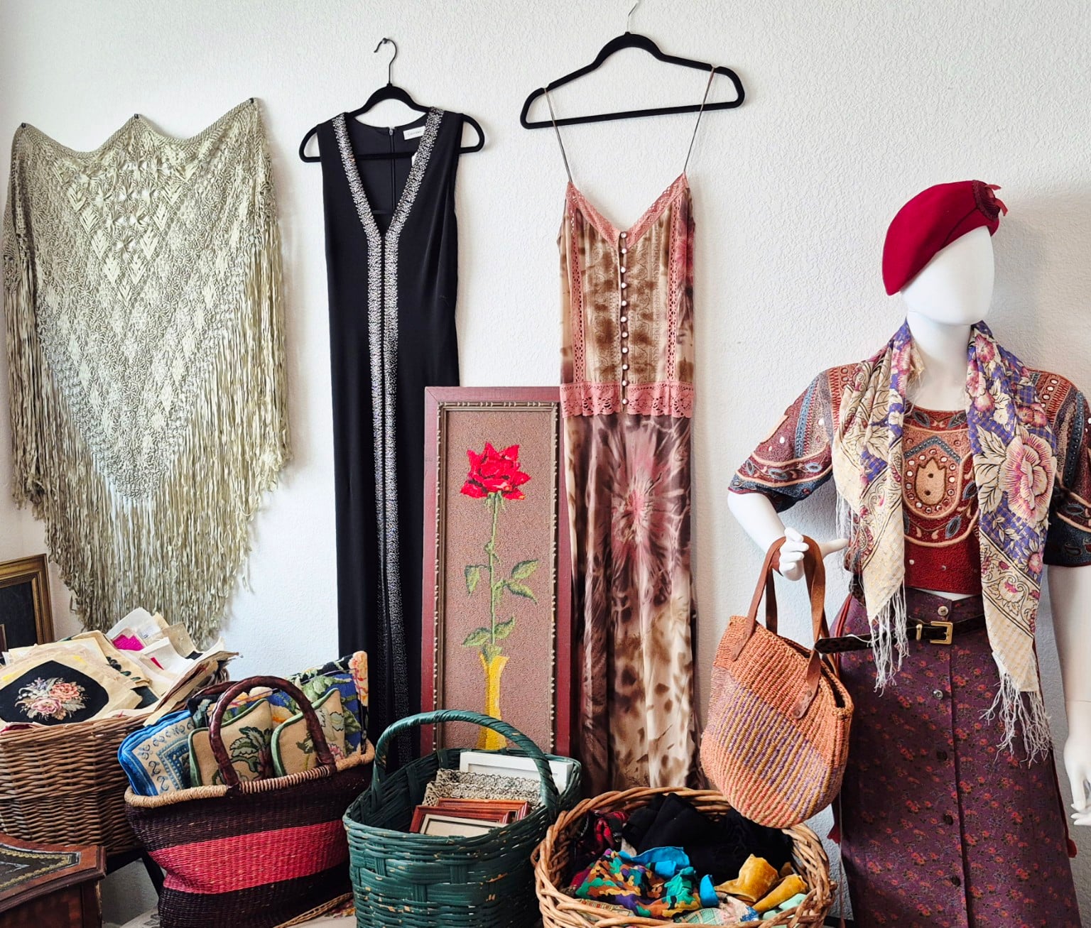 A sunlit corner of the shop with hanging dresses, a needlepoint rose and woven baskets